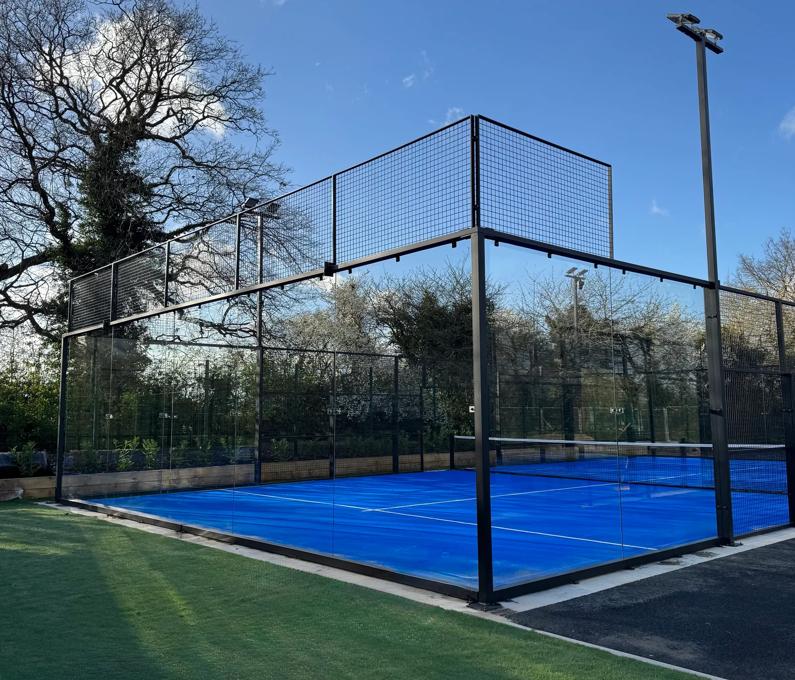 Blue outdoor padel court surrounded by glass, fencing and trees