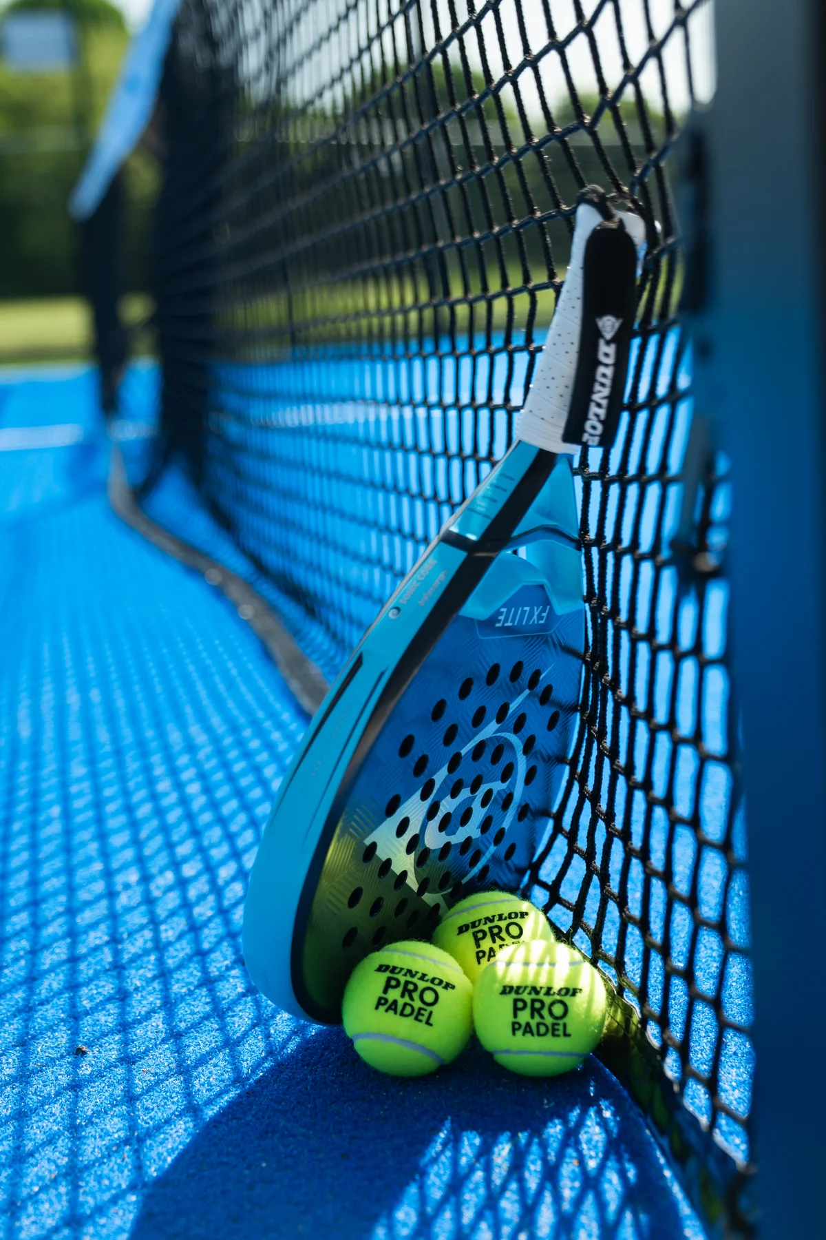 A padel racket and two yellow balls beside the net on a blue court